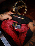 A close-up of a person's tattooed hands holding open a black quilted jacket or bag, revealing a bright red paisley-patterned interior lining. A Derby label is visible inside, and a gold chain handle is partially visible above.